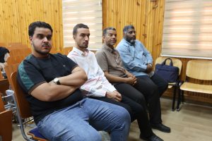 93115E6D-8D62-4AFE-8EF2-87BF6CD70EC0 - جامعة سبها | Sebha University Four men sit in a row of wooden chairs in a wood-paneled room, facing forward as if attending a lecture or meeting.