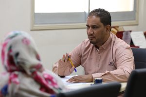 Man in a pink shirt explains something to a colleague across a desk, gesturing with a yellow pencil over papers in an office setting.