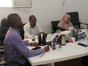 Three people sit at a white conference table with snacks and drinks in a casual meeting room.