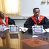 Two men in black robes with red sashes sit at a conference table with name placards and papers in front of them.