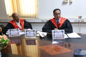 Two men in black robes with red sashes sit at a conference table with name placards and papers in front of them.