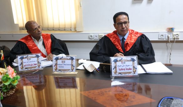 Two men in black robes with red sashes sit at a conference table with name placards and papers in front of them.