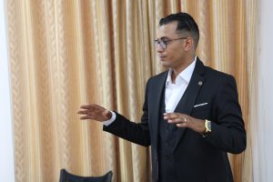 9A20F796-E05E-4BB1-924A-CABAD4316684 - جامعة سبها | Sebha University Man in a dark suit and white shirt gesturing with his hands while speaking, in front of gold patterned curtains.