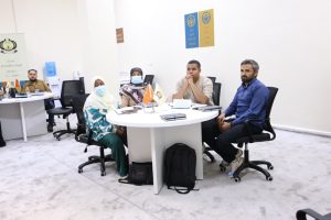 Four people sit around a white round table in a bright room, two women wearing headscarves and masks and two men, with notebooks and two small flags on the table.