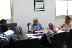 Group of adults seated around a conference table, discussing documents with notebooks and cups on the table; a framed certificate hangs on the wall behind them.