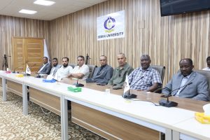 Group of men in suits seated along a long conference table in a meeting room, with a Sebha University banner on the wood-paneled wall behind them.