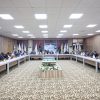 Large conference room with a U-shaped table setup and many attendees seated around it, flags along the back wall, and a decorative carpet.