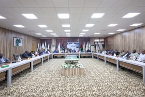 Large conference room with a U-shaped table setup and many attendees seated around it, flags along the back wall, and a decorative carpet.