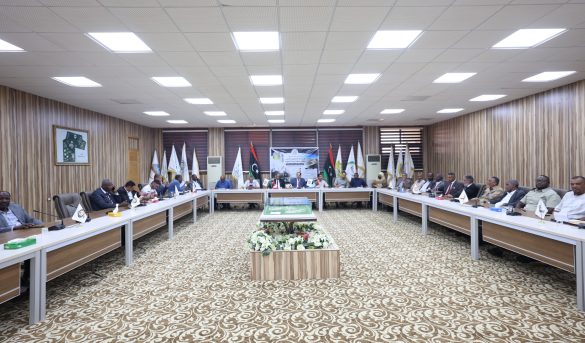 Large conference room with a U-shaped table setup and many attendees seated around it, flags along the back wall, and a decorative carpet.