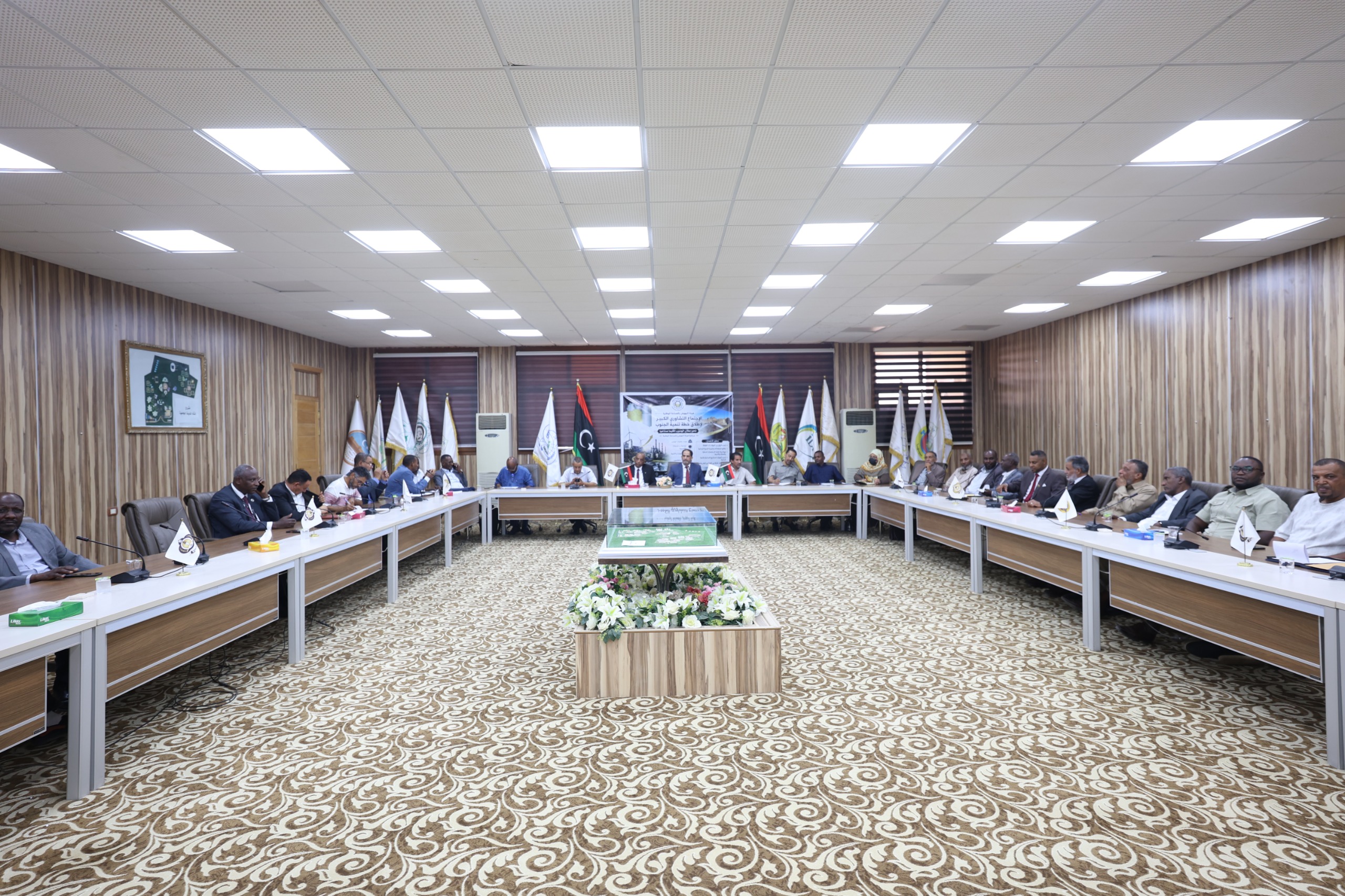 Large conference room with a U-shaped table setup and many attendees seated around it, flags along the back wall, and a decorative carpet.