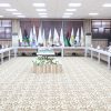 Large conference room with people seated around a U-shaped table, flags along the back wall, and a central floral centerpiece.