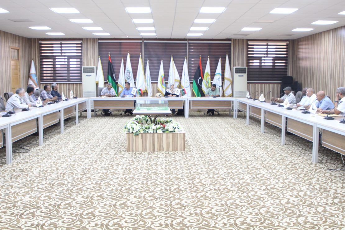 Large conference room with people seated around a U-shaped table, flags along the back wall, and a central floral centerpiece.