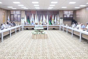 Large conference room with people seated around a U-shaped table, flags along the back wall, and a central floral centerpiece.