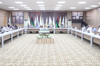 Large conference room with people seated around a U-shaped table, flags along the back wall, and a central floral centerpiece.