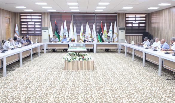 Large conference room with people seated around a U-shaped table, flags along the back wall, and a central floral centerpiece.