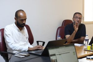 A273F3C2-C5F9-43AE-9F8B-7196423BD104 - جامعة سبها | Sebha University Two men sit at a conference table with laptops; one in a white shirt looks down while the other in a dark polo speaks and gestures with his hand.