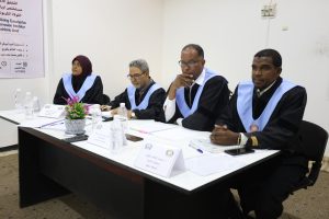 A3FD156A-6721-457C-A588-E15AA13F4360 - جامعة سبها | Sebha University Four graduates in black robes with blue stoles sit at a table for a panel, with name cards and water bottles on the table.