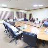 Group of people seated around a large conference table in a meeting room, discussing or reviewing documents.