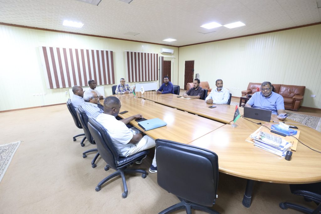 A56208B6-2746-4127-A04C-C8C9F051BD38 - جامعة سبها | Sebha University Group of people seated around a large conference table in a meeting room, discussing or reviewing documents.