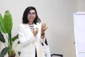 Woman in a white blazer giving a presentation, gesturing with hands in a bright room with a large plant nearby.