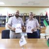 Two men in a formal office hold documents with a logo, standing behind a conference table with Libyan flags on the table.
