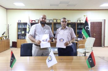 Two men in a formal office hold documents with a logo, standing behind a conference table with Libyan flags on the table.