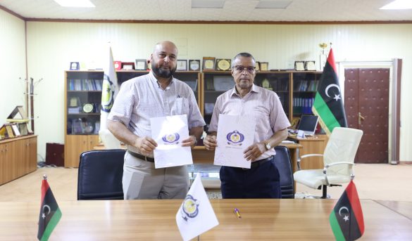 Two men in a formal office hold documents with a logo, standing behind a conference table with Libyan flags on the table.