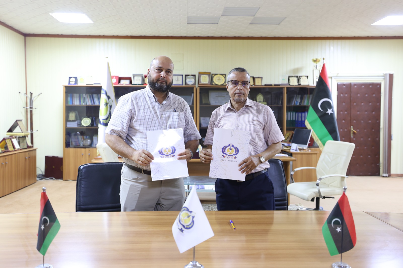 Two men in a formal office hold documents with a logo, standing behind a conference table with Libyan flags on the table.