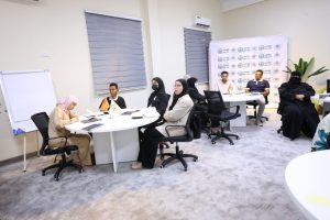 Group of people in a training room seated around round tables, some in hijabs, with a flip chart and a logo banner in the background.