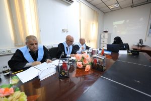 Four panelists sit at a curved conference table; three men in black robes with light blue sashes and a woman in a hijab uses a laptop, with a projected presentation on the wall behind them