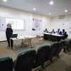 Lecturer at a desk presenting a slideshows to a classroom, with a projection screen showing a slide; five panelists in gowns sit at a table on the right.