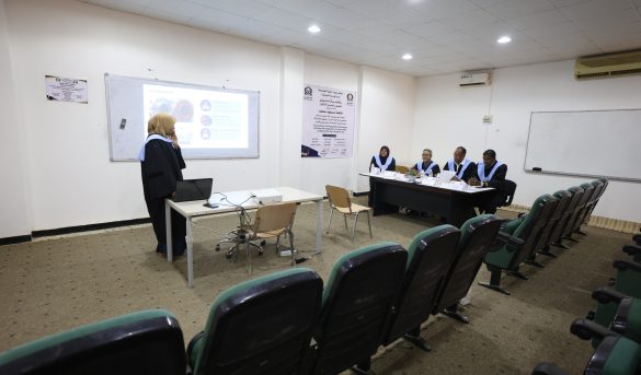 Lecturer at a desk presenting a slideshows to a classroom, with a projection screen showing a slide; five panelists in gowns sit at a table on the right.