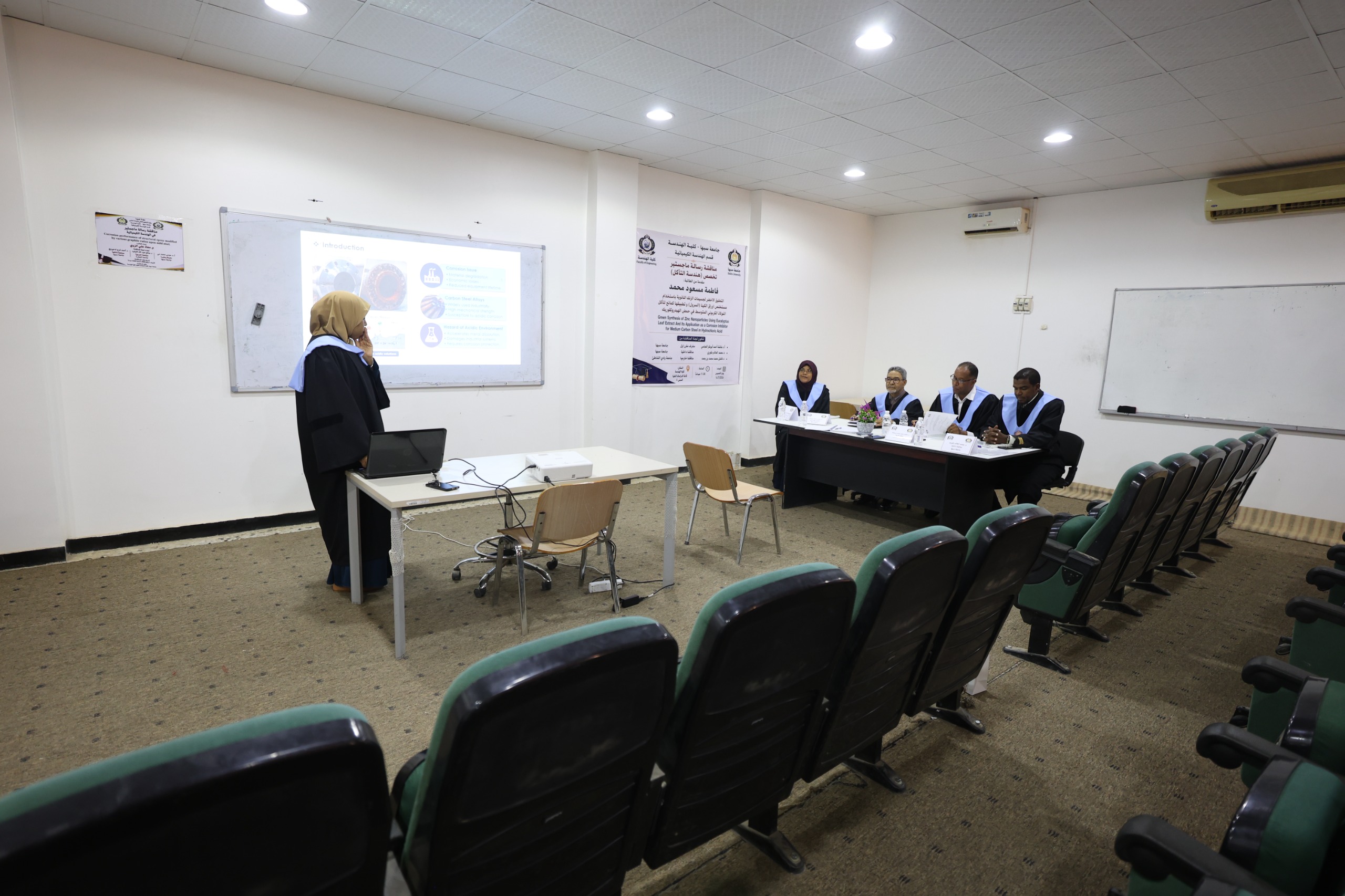 Lecturer at a desk presenting a slideshows to a classroom, with a projection screen showing a slide; five panelists in gowns sit at a table on the right.