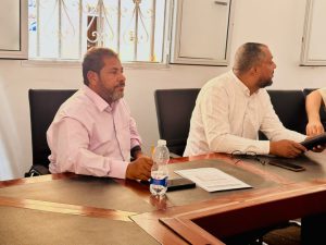 A9FE445C-2ACE-4AA4-94F9-B90FAB0733F8 - جامعة سبها | Sebha University Two men sit at a conference table in a bright room, facing to the right; one wears a pink shirt, the other a light-colored shirt, with papers and a water bottle on the table in front of them.