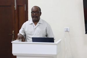 Middle-aged man wearing glasses stands at a white lectern with a laptop, delivering a speech in a room.