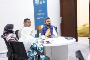 Group of people in a round-table discussion wearing masks in a bright room, with a blue Arabic banner in the background.