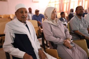 An elderly man in a white cap and robe sits beside a woman in a light hijab at a conference room audience. In the background, other attendees listen attentively.