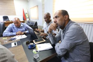 AE868F9A-8359-4042-8D65-8DF2632B28F9 - جامعة سبها | Sebha University Three men in a meeting room sit around a conference table with laptops and papers, discussing work.