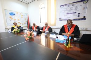 Four academics in black robes with red sashes sit at a conference table during a formal meeting, with laptops, flowers, and a flag on the table and a poster on the wall behind them.