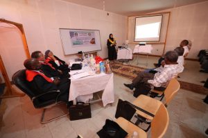 Speaker in a yellow scarf presents to a group seated around a table in a classroom with a projector screen at the front.