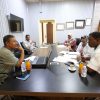 Five people sit around a conference table in a meeting, with papers, a laptop, and drinks on the table./nMeeting scene in an office with documents and beverages nearby.