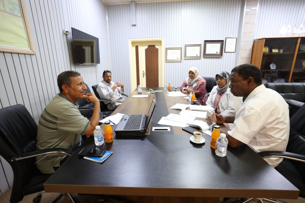 Five people sit around a conference table in a meeting, with papers, a laptop, and drinks on the table./nMeeting scene in an office with documents and beverages nearby.
