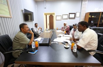 Five people sit around a conference table in a meeting, with papers, a laptop, and drinks on the table./nMeeting scene in an office with documents and beverages nearby.