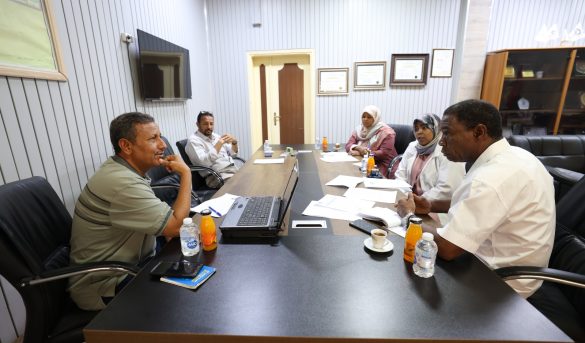B6C8641B-1724-474F-B890-F1659EF08C01 - جامعة سبها | Sebha University Five people sit around a conference table in a meeting, with papers, a laptop, and drinks on the table./nMeeting scene in an office with documents and beverages nearby.