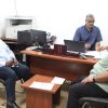 Three men sit around a wooden desk in an office, discussing documents and working on laptops.