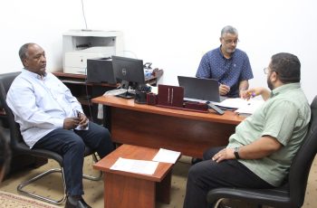 Three men sit around a wooden desk in an office, discussing documents and working on laptops.