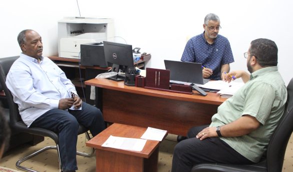 Three men sit around a wooden desk in an office, discussing documents and working on laptops.