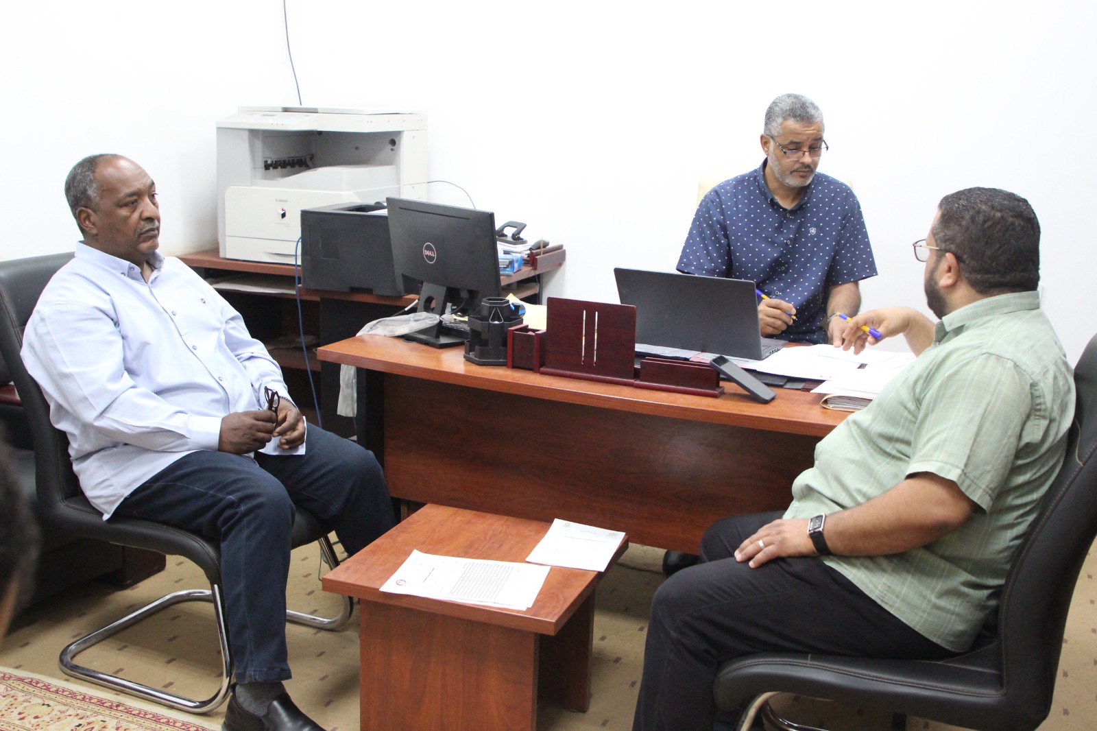Three men sit around a wooden desk in an office, discussing documents and working on laptops.