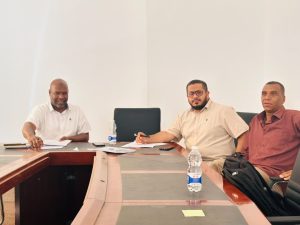 BA55DE56-84B2-4E1E-87A2-495F9E59F5FB - جامعة سبها | Sebha University Three men sit at a conference table with papers and a laptop, engaged in a business meeting.