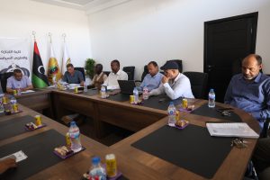 BA90AA11-A92B-4F85-BB15-2EC707A2E706 - جامعة سبها | Sebha University Group of people in a formal meeting around a long conference table with flags and a banner in the background.